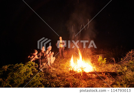 Young family drinking tea next to fire place. Cozy night in forest. Family of four warming near campfire. Young family drinking tea next to fire place. Cozy night in forest. Family of four warming near campfire. 114793013