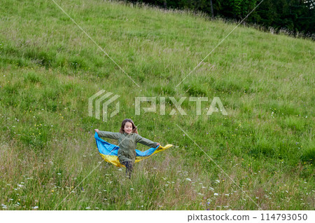 Happy Ukrainian running in Carpathian mountains. Little kid smiling and flattering Ukrainian flag on background of hills at sunset. 114793050