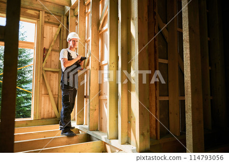 Carpenter constructing wooden framed house. Man worker working with screwdriver, wearing work overalls and helmet. Concept of modern eco-friendly construction. 114793056