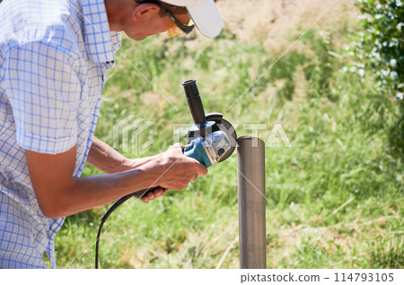 Male worker cutting metal pole with electric circular saw. Man in protective glasses using hand-held grinder while preparing aluminum pole for solar panel installation. Male worker cutting metal pole with electric circular saw. Man in protective glasses using hand-held grinder while preparing aluminum pole for solar panel installation. 114793105