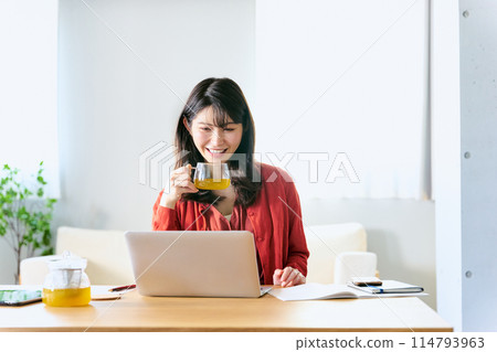 A Japanese woman in her 30s working remotely on a laptop at home with a bright smile while drinking tea 114793963