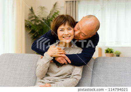 一位年長的丈夫在家裡客廳的沙發上從後面擁抱並親吻妻子，以表達他的愛。 114794021