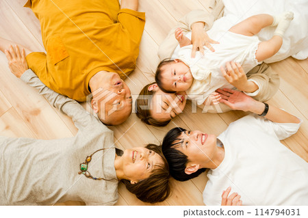 A family of five people, three generations of parents and grandparents, including a 0-year-old baby, lying in a circle on the floor on their backs. 114794031