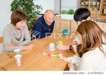 A baby girl reaches for a toy on a desk in the living room at home while her mother, father, grandfather and grandmother watch her 114794083