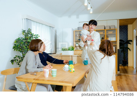 A morning or early morning scene of a family of five in the living room at home, with a baby being held by the father and the mother, grandfather, and grandmother watching over them 114794086