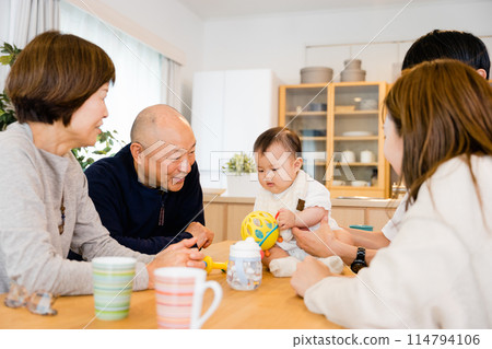 A smiling family of 5 people from 3 generations including senior grandparents and parents holding and soothing a baby. 114794106