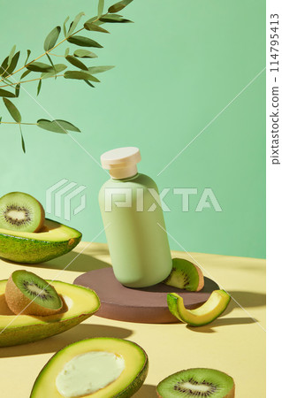 A cosmetic bottle without label displayed on a brown pedestal with a kiwi piece decorated, still has blank space for adding designing elements, surrounded by fresh fruits flat lay on yellow table top 114795413