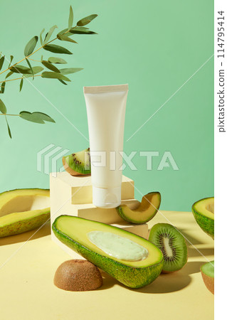Vertical photo on light green background, a ladder-shaped platform with fresh avocado, kiwi scattered lay and a blank cosmetic tube displayed above. Mock up for advertising with space for text 114795414