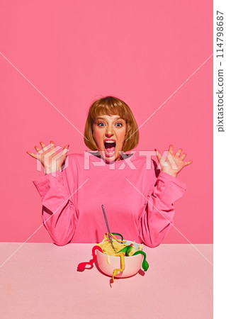 Surprise in Bowl. Yong woman in pink sweatshirt, expressing shock, shouting as founds in a bowl of spaghetti jelly snakes. Pink background. Surprise in Bowl. Yong woman in pink sweatshirt, expressing shock, shouting as founds in a bowl of spaghetti jelly snakes. Pink background. 114798687