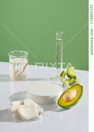 Creative photo was shot from above against green background, laboratory items, avocado and coconut displayed on white table and a blank round pedestal. Blank space for showing product or adding text 114802325