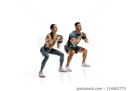 Man and woman exercising with kettlebells in squat position against white studio background. Athletic movements. Man and woman exercising with kettlebells in squat position against white studio background. Athletic movements. 114802775