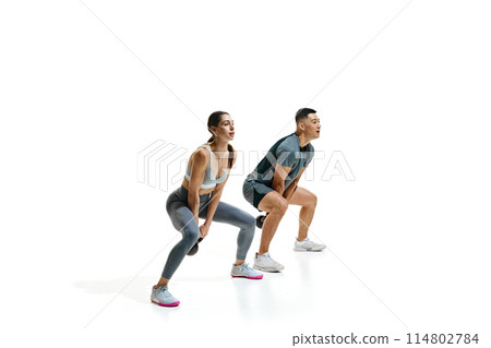 Young, fit man and woman captured mid-exercise, performing squats with kettlebells against white studio background. 114802784