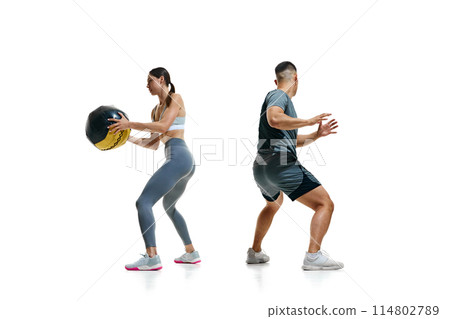 Pair of athletes engaged in dynamic exercise involving large black medicine ball against white studio background. 114802789
