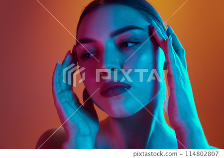 Close-up of young woman gently touching the sides of her face in neon light against gradient background. 114802807