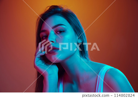 Tender young woman with long, dark hair posing with her head resting on her hand in neon light against gradient background. 114802824