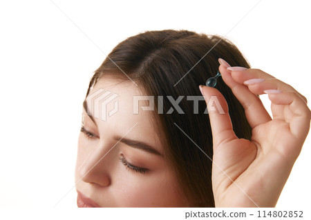 Close up of young brunette woman's head with silky, straight hair holding capsule with oil against white background. Close up of young brunette woman's head with silky, straight hair holding capsule with oil against white background. 114802852