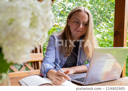 Young happy woman focuses on her laptop in wooden alcove. Relaxed outdoor setting emphasizes comfort and productivity. Remote work learning Young happy woman focuses on her laptop in wooden alcove. Relaxed outdoor setting emphasizes comfort and productivity. Remote work learning 114802985