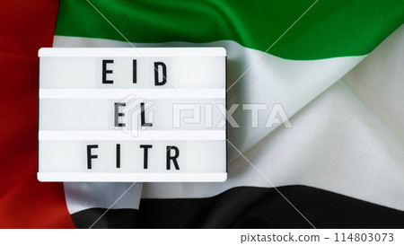 EID EL FITR - Eid Mubarak - Happy Holidays text frame on United Arab Emirates waving flag made from silk material. Public holiday celebration background. The National Flag of UAE. Commemoration Day 114803073