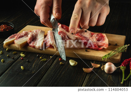 Cutting lard with a knife in a man's hand to prepare a delicious snack on the kitchen table with pepper and garlic. Peasant food concept 114804053