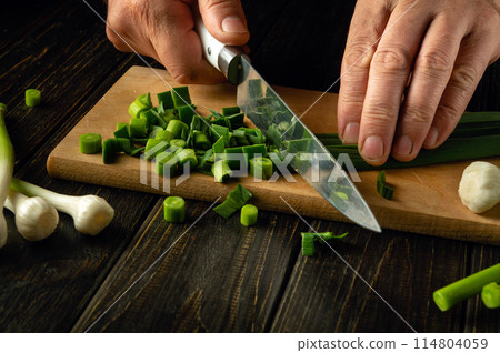 The chef cuts green young garlic on a wooden cutting board with a knife for preparing vegetarian food. Advertising space 114804059