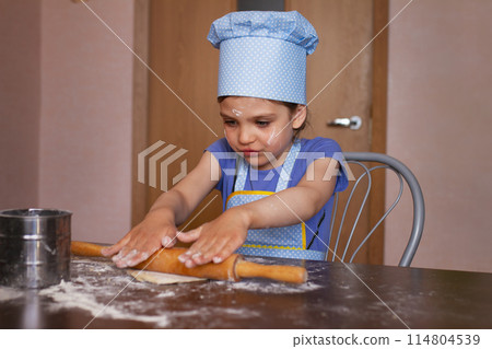 Portrait of 5 year old girl in blue dress and dressed as chef in kitchen laughs and rolling out dough Portrait of 5 year old girl in blue dress and dressed as chef in kitchen laughs and rolling out dough 114804539