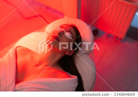 Top view face of female client lying during non-invasive type of phototherapy. Closeup of woman patient in protective goggles undergoes blue LED light therapy for skin rejuvenation at aesthetic clinic 114804609