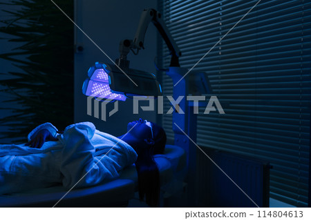 Remote view of female patient undergoes blue LED light therapy for skin rejuvenation at modern aesthetic clinic. Using phototherapy device with blue light wavelength for acne treatments. Remote view of female patient undergoes blue LED light therapy for skin rejuvenation at modern aesthetic clinic. Using phototherapy device with blue light wavelength for acne treatments. 114804613