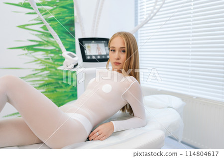 Indoors shot of sensual blonde female patient in white uniform lying on medical couch in modern clinic while waiting beautician specialist, looking at camera. Concept of spa, beauty treatment. 114804617