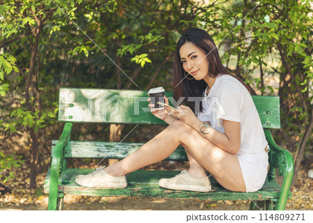 Asian Woman use smartphone drinking coffee hand holding hot disposable cup in green park. Banner Happy Relax asian woman smiling face at outdoors garden. Young women enjoy nature with copy space Asian Woman use smartphone drinking coffee hand holding hot disposable cup in green park. Banner Happy Relax asian woman smiling face at outdoors garden. Young women enjoy nature with copy space 114809271
