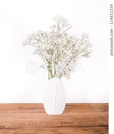 There is a white vase containing flowers placed on a wooden table 114811334