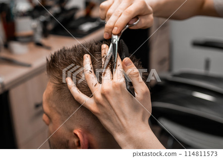 Professional barber styling hair of his client in the barbershop using scissors and comb. 114811573