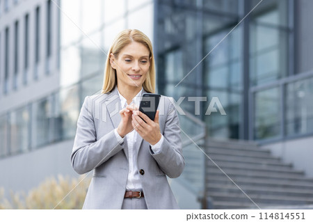 A professional woman in a gray suit smiles as she uses a smartphone outside a modern office building. Emphasizing connectivity and business efficiency. 114814551