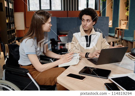 High angle view of Caucasian girl with disability and her Middle Eastern friend sitting at table in school library working together on presentation High angle view of Caucasian girl with disability and her Middle Eastern friend sitting at table in school library working together on presentation 114819294