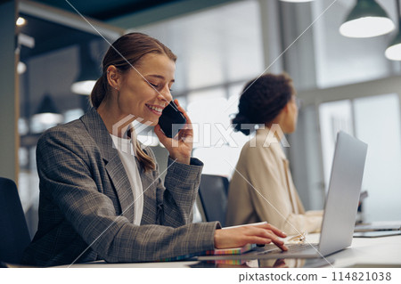 Woman sales manager talking by phone with client sitting in modern coworking and working on laptop Woman sales manager talking by phone with client sitting in modern coworking and working on laptop 114821038