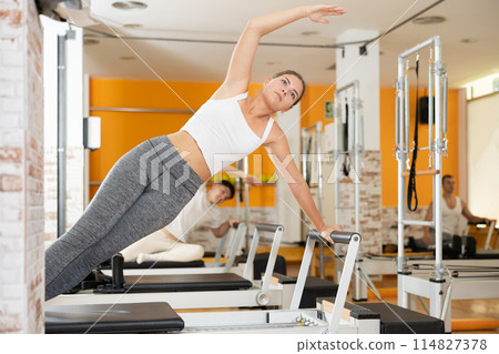 Girl performing side plank with arm extended on Pilates reformer 114827378