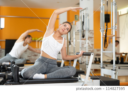 Girl doing side stretching with arm extended on Pilates reformer Girl doing side stretching with arm extended on Pilates reformer 114827395