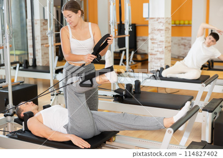Man athlete performs exercises to increase leg strength using sling reformer under coach guidance. Man athlete performs exercises to increase leg strength using sling reformer under coach guidance. 114827402