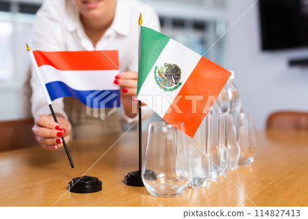 Preparing for business negotiations - woman sets small flags of countries of Netherlands and Mexico on table 114827413