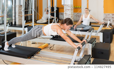 Girl performing exercises on Pilates reformer in fitness studio Girl performing exercises on Pilates reformer in fitness studio 114827495