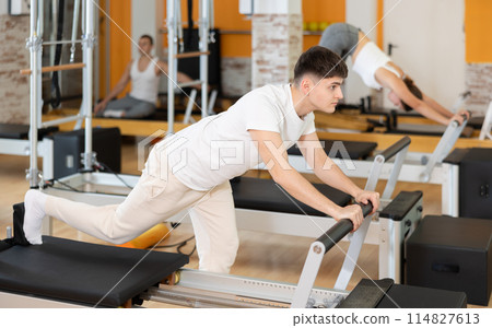 Man works out in gym and performs set of exercises to stretch leg muscles with help of reformer 114827613