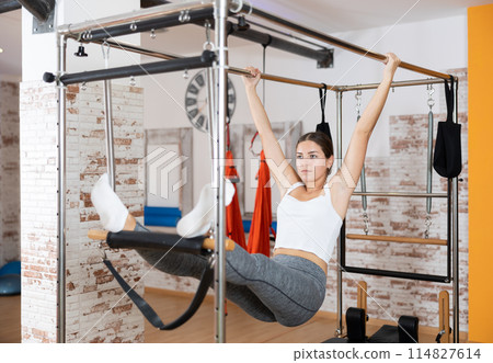 Girl visitor performs exercises in hanging position on combined Pilates trapeze table. Girl visitor performs exercises in hanging position on combined Pilates trapeze table. 114827614
