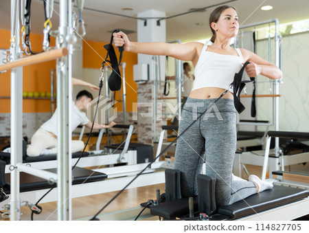 Girl in gym with help of rope reformer performs exercises to strengthen muscles of her arms. 114827705