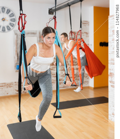 Girl visitor performs exercises in hanging position on Pilates slings hammock Girl visitor performs exercises in hanging position on Pilates slings hammock 114827968