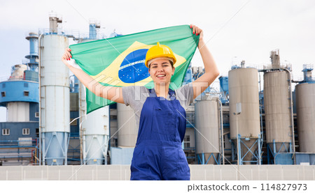 Young female engineer in helmet waving state flag of Brazil while standing in front of big tanks at chemical plant 114827973