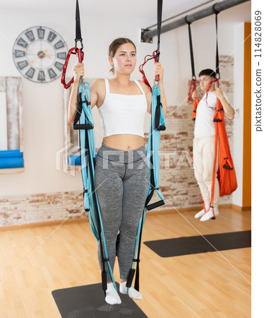 Girl visitor performs exercises in hanging position on Pilates slings hammock 114828069