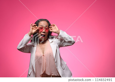 Surprised elderly female in sunglasses and stylish clothes posing over pink background with copy space 114832491