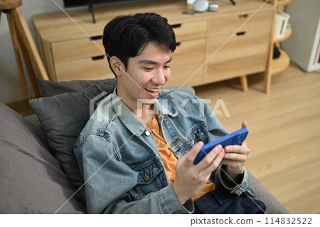 Smiling man playing video game on smart phone while sitting on couch at home Smiling man playing video game on smart phone while sitting on couch at home 114832522
