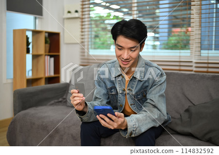Smiling young Asian man doing online payment through credit card and smart phone Smiling young Asian man doing online payment through credit card and smart phone 114832592