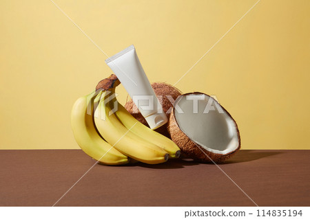 An unlabeled cosmetic tube is leaning on a bunch of fresh banana and a few dried coconuts on a brown table top against yellow texture. Photo for advertising with front shot and copy space 114835194
