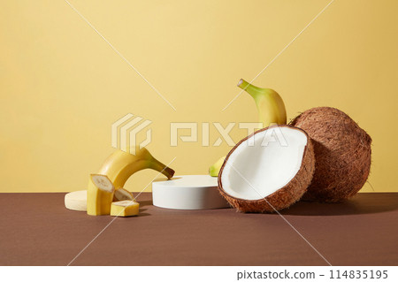 An white empty showcase podium in circular shaped placed in center next to fresh yellow bananas, coconuts and some slices of banana decorated around. Front view photo with blank space for design 114835195